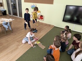 Dzieci młodsze siedzą na dywanie. Przed nimi stoi troje starszych dzieci, które pokazują młodszym kolegom i koleżankom swoje wytwory pracy zrobione z kartonów i innych materiałów nadających się do recyklingu.