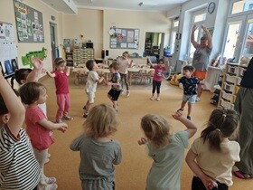 12 dzieci i dwie kobiety stoją w kole w sali przedszkolnej i tańczą. W dalszej części sali stoją stoliki dziecięce i krzesełka.