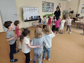 12 dzieci i 1 kobieta stoją w sali na dywanie: 7 dzieci stoi blisko kobiety, 5 jest oddalonych. Przy ścianie stoi biała tablica, na której przywieszone są kolorowe ilustracje.