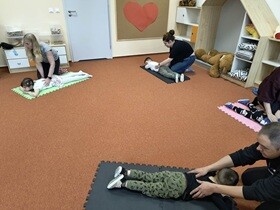 Na rozłożonych matach leży czwórka dzieci. Obok każdego dziecka jest dorosły, który masuje plecy swojego dziecka.