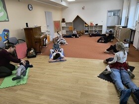 Na sali, na kolorowych matach leży ośmioro dorosłych. Część dzieci siedzi na plecach rodziców, dwoje dzieci siedzi obok. Wszystkie dzieci masują plecy swojego rodzica.