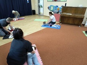 Troje dzieci leży na matach, dorośli je masują. Dorośli patrzą w stronę osoby z jasnymi włosami, która masuje dziewczynkę.