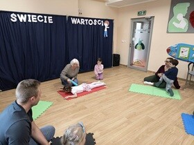 Na czerwonej macie leży dziewczynka na plecach. Kobieta siedząca obok trzyma w dłoni niebieski wałek, którym dotyka plecy dziecka.