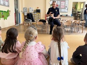 Na dywanie siedzą dzieci. Przed nimi na krześle siedzi pan w mundurze strażnika miejskiego, na kolanach trzyma pluszaka Rysia.