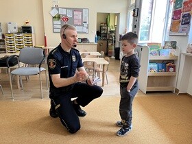 Strażnik miejski kuca przed chłopcem. Pan w jednej dłoni trzyma długopis a w drugiej notes.