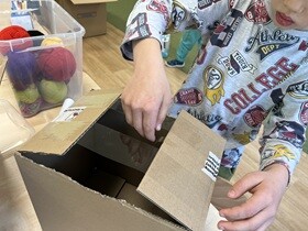 Chłopiec ma przed sobą karton, do którego przykleja taśmę przezroczystą.