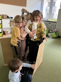 Na dywanie ustawiony jest duży karton, w którym stoją dzieci. Przed kartonem na dywanie siedzi chłopiec w niebieskiej bluzce.