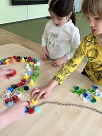 Dwójka dzieci stoi przy stole i układa zakrętki wzdłuż sznurka.