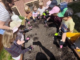 Dzieci stoją naokoło drewnianej skrzyni wypełnionej ziemią. Przedszkolaki trzymają w rękach różowe i zielone łopatki, którymi przekopują ziemię.