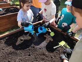 Czworo dzieci stoi naokoło drewnianej skrzyni wypełnionej ziemią. Dzieci mają nałożone rękawiczki i w rękach trzymają małe konewki, którymi podlewają ziemię.