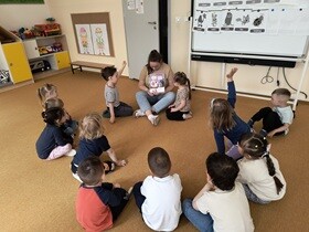 Dzieci wraz z nauczycielką siedzą na dywanie. Nauczycielka pokazuje dzieciom stronę w książce o emocjach, na której zilustrowany jest strach. Dwoje dzieci trzyma ręce uniesione w górę.