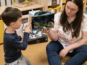 Nauczycielka i chłopiec siedzą na dywanie. Nauczycielka trzyma w rękach makietę akwarium a chłopiec pokazuje na wybrane elementy i opowiada o nich.