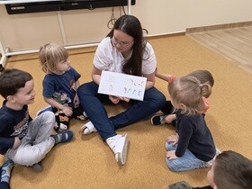 Nauczycielka z dziećmi siedzi na dywanie. Prowadząca pokazuje dzieciom książkę, której głównym bohaterem jest jednorożec Gucio.
