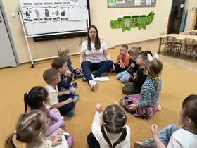 Nauczycielka z dziećmi siedzą w kole na dywanie. Razem wykonują ćwiczenie oddechowe. Wszyscy mają zamknięte oczy i opuszczone ręce z dłońmi zaciśniętymi w pięściach.