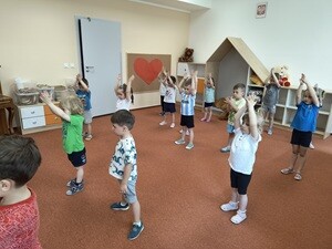 Dzieci stoją w rozsypce na dywanie, tańczą i trzymają złączone ręce uniesione w górę. Dzieci ubrane są w stroje gimnastyczne.