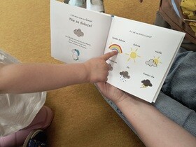 Nauczycielka trzyma książkę z ilustracjami przedstawiającymi Gucia-jednorożca oraz ikonami tęczy, słońca, burzy, chmur, które pomagają wyrazić obecny nastrój. Dziecko wskazuje palcem na swój obecny nastrój oznaczony jako tęcza.