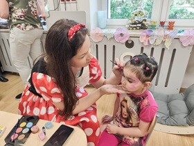 Kobieta w ubraniu Myszki Miki maluje twarz dziewczynce w różowej bluzeczce. Obie siedzą na krzesełkach przy stoliku.