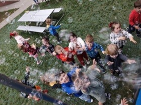 Dzieci stoją na trawie na placu zabaw i łapią bańki mydlane, lecące na nie z góry.