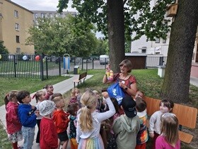 Grupka dzieci stoi na placu zabaw zwrócona twarzą do kobiety w kolorowej sukience, która mówi do dzieci trzymając w ręce uniesionej do góry kolorowe foremki do zabawy w piaskownicy.
