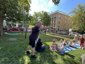 Na kocach siedzą dzieci, z lewej strony stoi pan ubrany na czarno z wyciągniętą ręką w stronę widowni.
