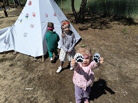 Troje dzieci znajduje się przed białym namiotem w stylu tipi. Jeden chłopiec wchodzi do namiotu. Dziewczynka trzyma uniesione, wydrukowane ślady zwierzęcia.
