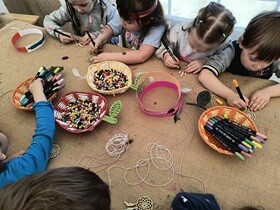 Dzieci siedzą przy stole i malują kolorowymi flamastrami drewniane medaliony. Na głowach mają samodzielnie zrobione, papierowe opaski w stylu indiańskim. Na stoliku stoją pojemniki z kolorowymi koralikami, flamastrami i sznurkami.