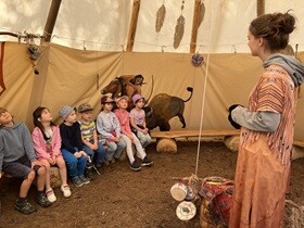 Siedmioro dzieci siedzi na ławce w namiocie, przed nimi stoi pani w stroju indiańskim. Po prawej stronie, na ścianie widać malowidło Indianina polującego na bizona.