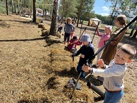 Dwóch chłopców próbuje naciągnąć łuk. Za nimi stoją pozostałe dzieci.